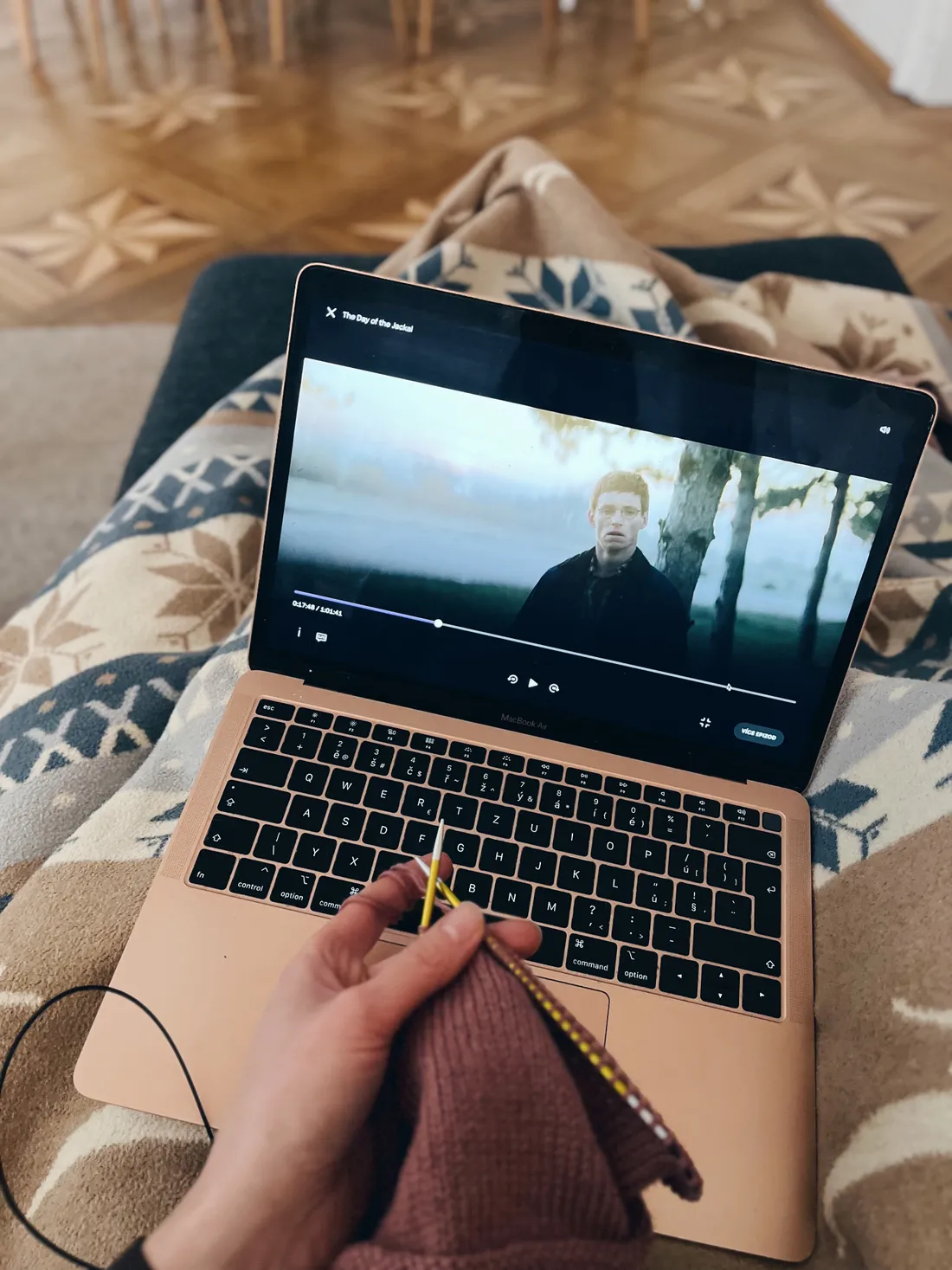 Růžový MacBook s filmem The Day of the Jackal na klíně, ruce pletou bordó svetr pod dekou.