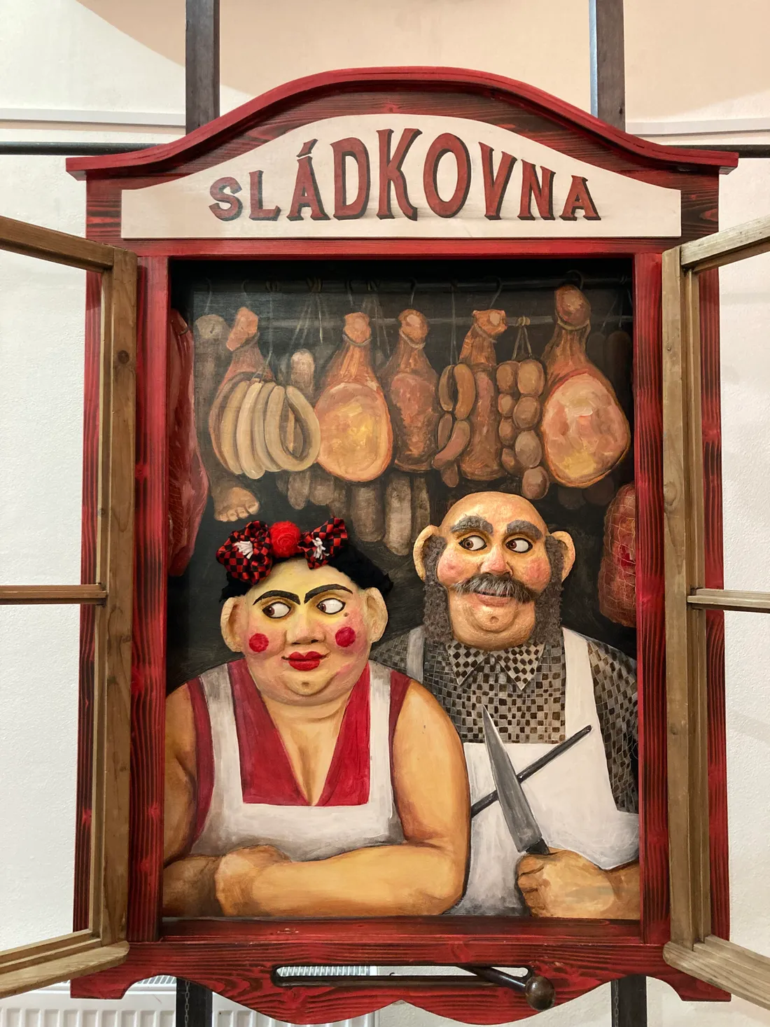 Loutková scéna SLÁDKOVNA s postavami prodavačů, noži a visícími uzeninami v červeném rámu výlohy.