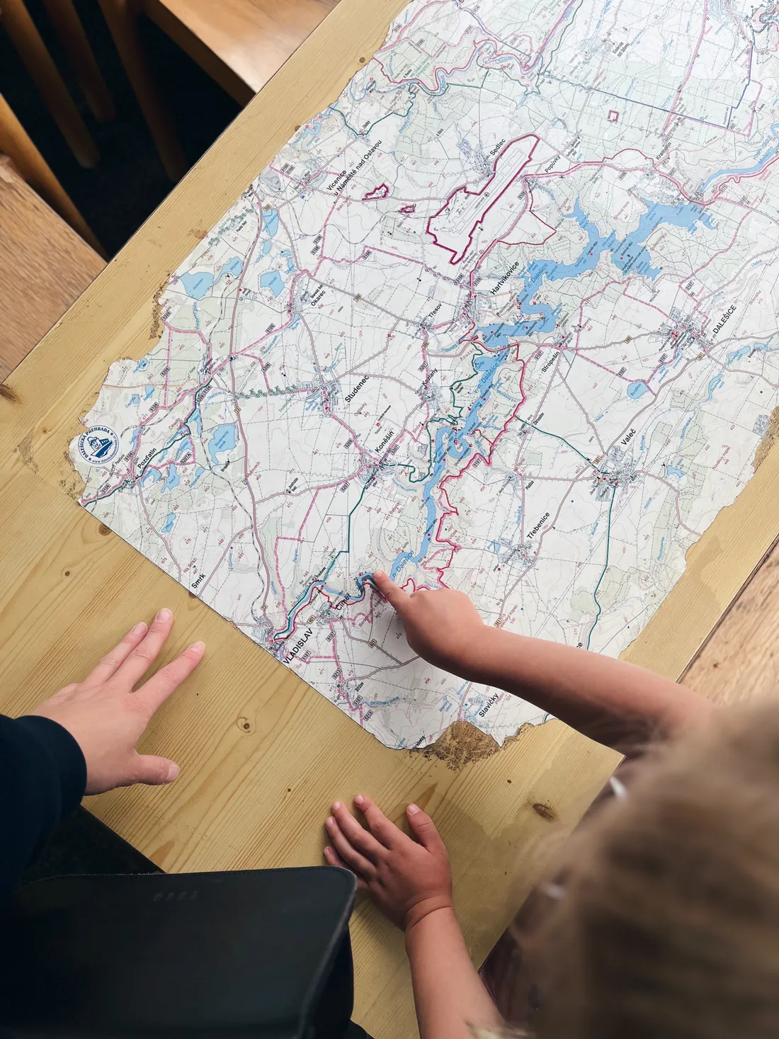 Dětská ruka ukazuje místo na turistické mapě Dalešické přehrady rozložené na dřevěném stole.