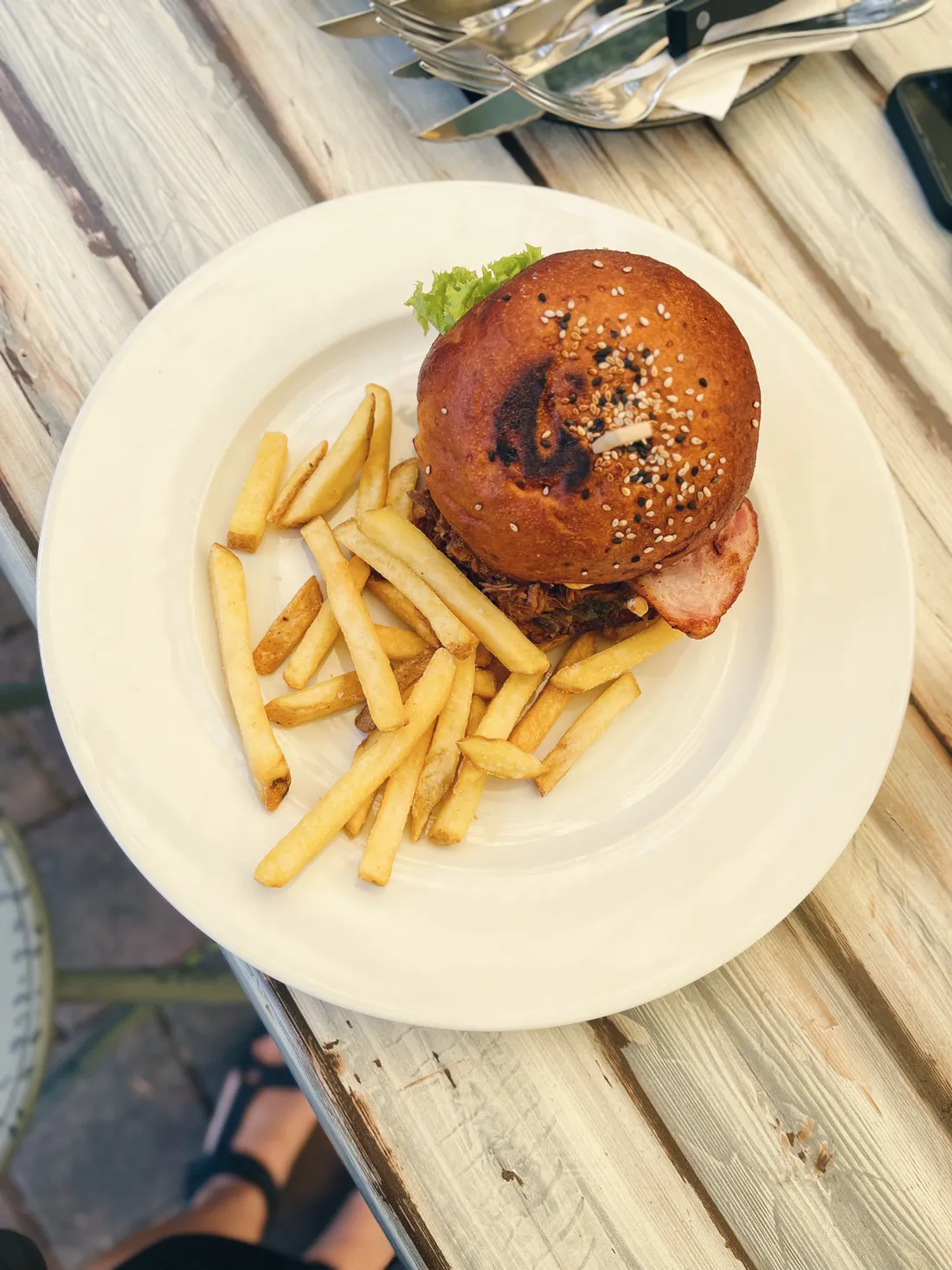Burger se slaninou a hranolky na bílém talíři na dřevěném stole venku.