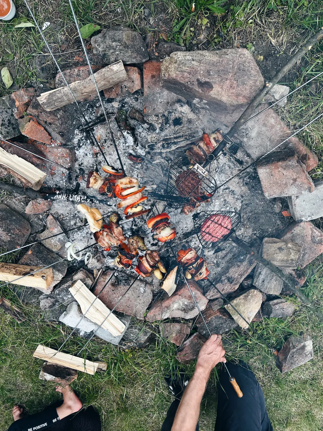 Pohled shora na kruhové ohniště z kamenů se špízy masem a zeleninou a mřížkou s placičkami nad žhavými uhlíky.