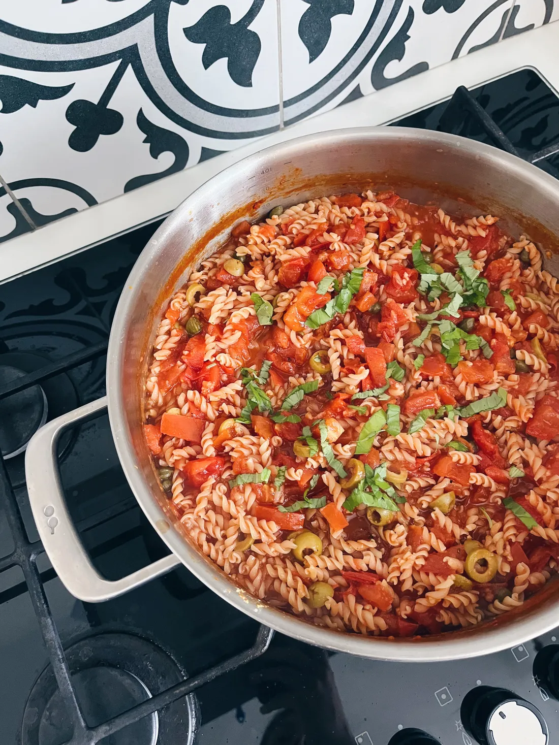 Pánev s těstovinami fusilli v rajčatové omáčce, olivami a bazalkou na černé plynové desce.
