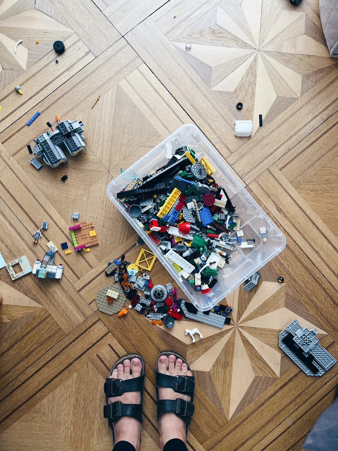 Průhledný box plný kostek LEGO a rozsypaných dílků na parketách s hvězdicovým vzorem, nohy v černých sandálech.