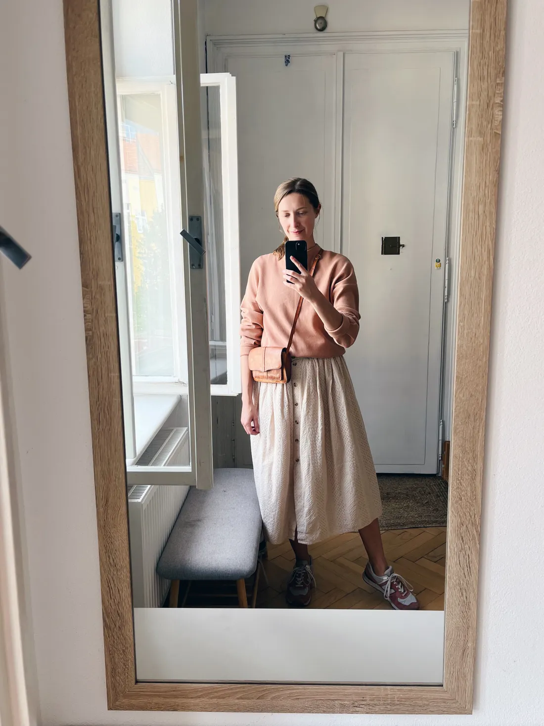 Selfie v zrcadle v broskvové mikině a bílé sukni s knoflíky a teniskami New Balance v chodbě.