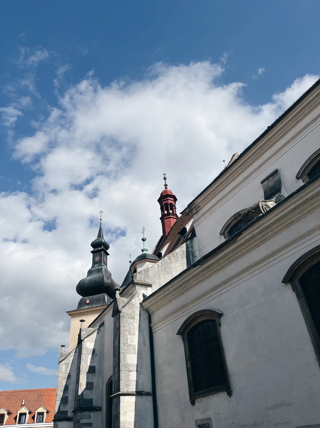 Bílá barokní budova s černou cibulovitou věží a červenou věží proti modré obloze.