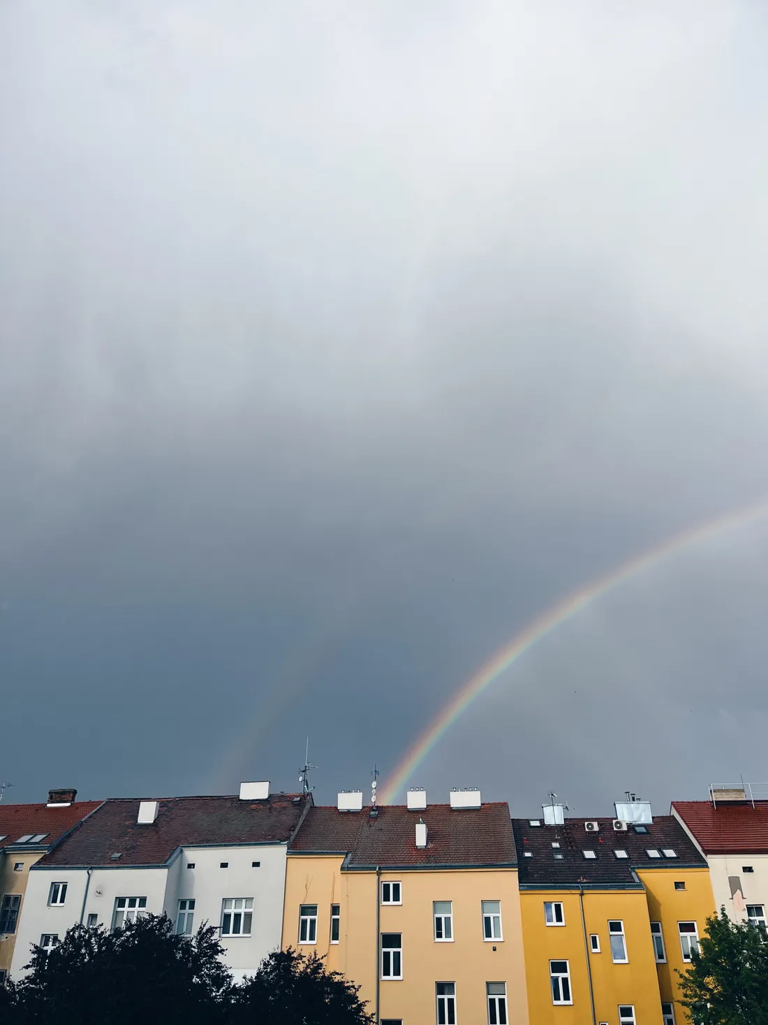 Dvojitá duha nad řadou barevných činžovních domů pod tmavými mraky.