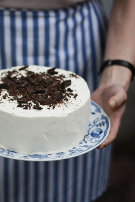 Bílý piškotový dort s mascarpone posypaný čokoládou, držený v rukou nad krajkovým talířem.
