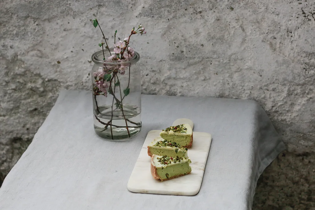 Tři řezy pistáciového cheesecaku na mramorovém prkně a váza s růžovými květy.