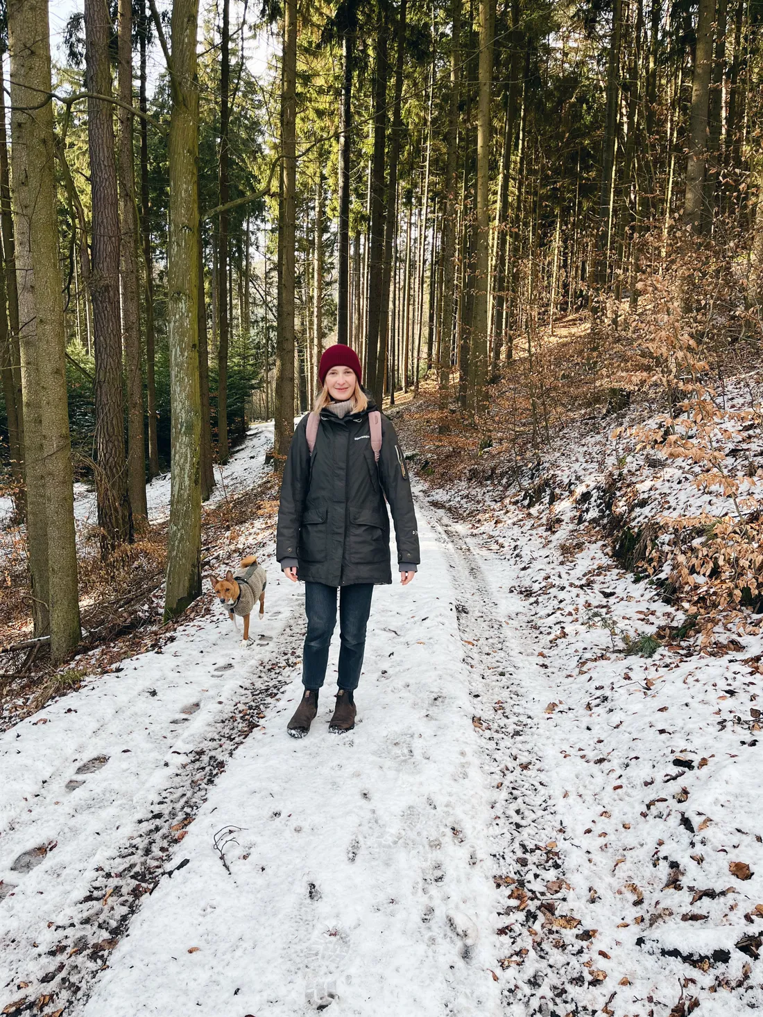 Žena v tmavé zimní bundě a červené čepici na zasněžené lesní cestě, vedle ní kráčí malý hnědý pes.