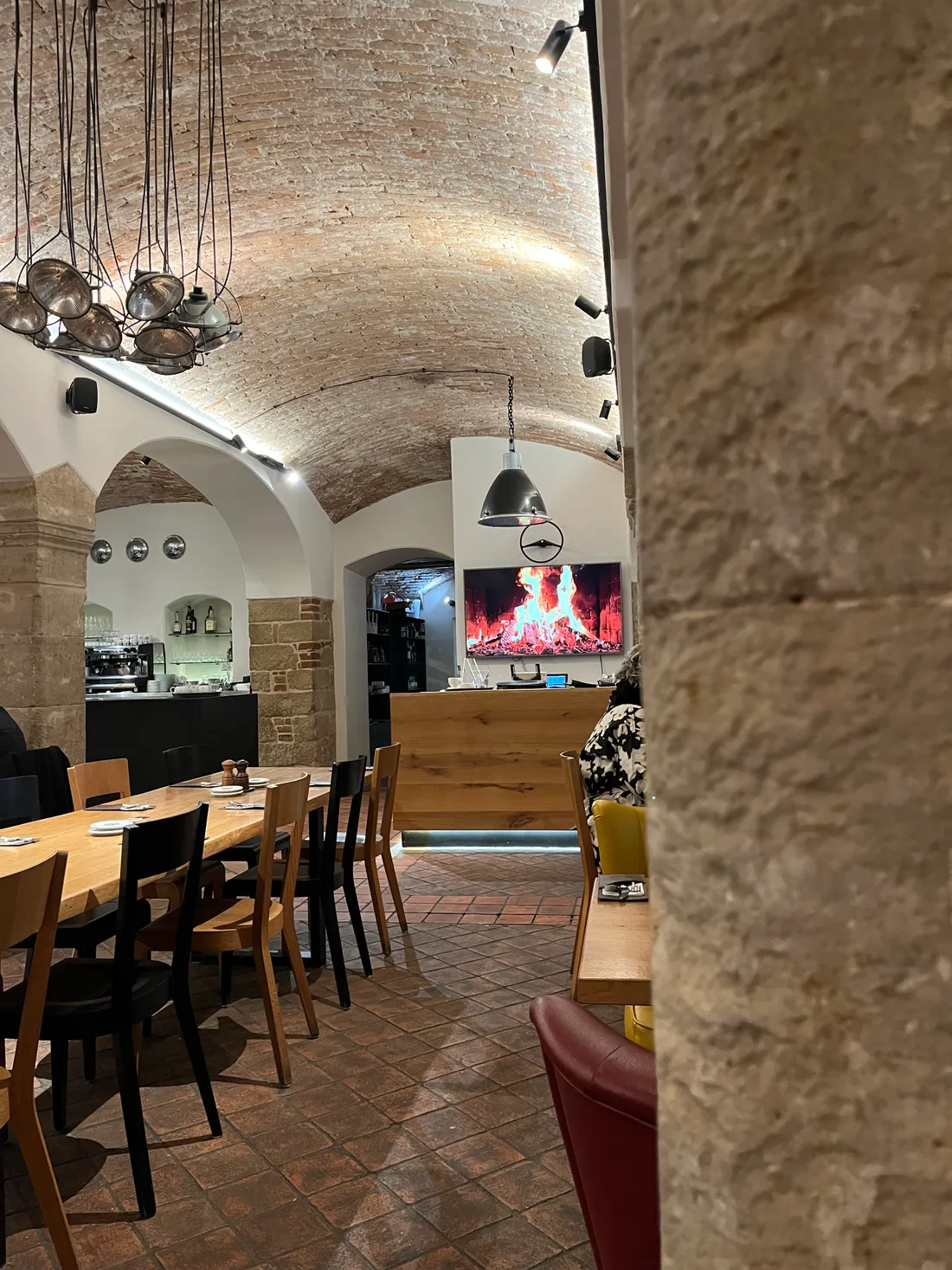 Interiér restaurace s klenutým cihlovým stropem, dlouhými stoly a obrazovkou s krbem nad dřevěným pultem.