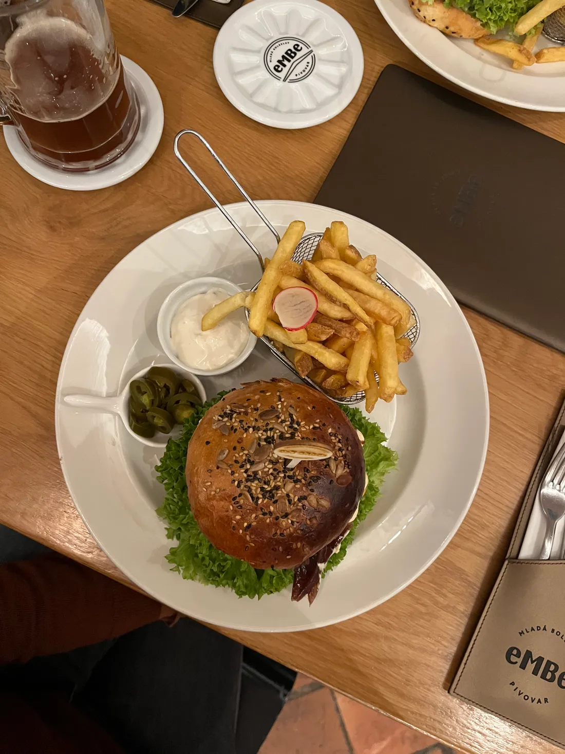 Burger se semínkovou bulkou, hranolky v košíku a omáčkami na bílém talíři s pivem na dřevěném stole.