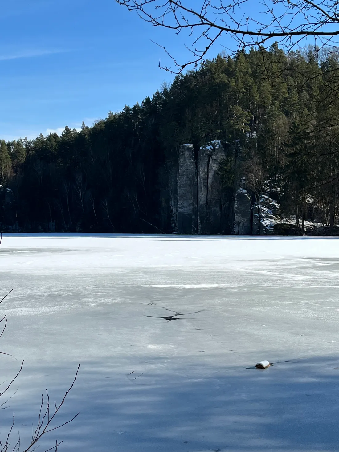 Zasněžené zamrzlé jezero s prasklinou v ledu, v pozadí skalní útvary a les.