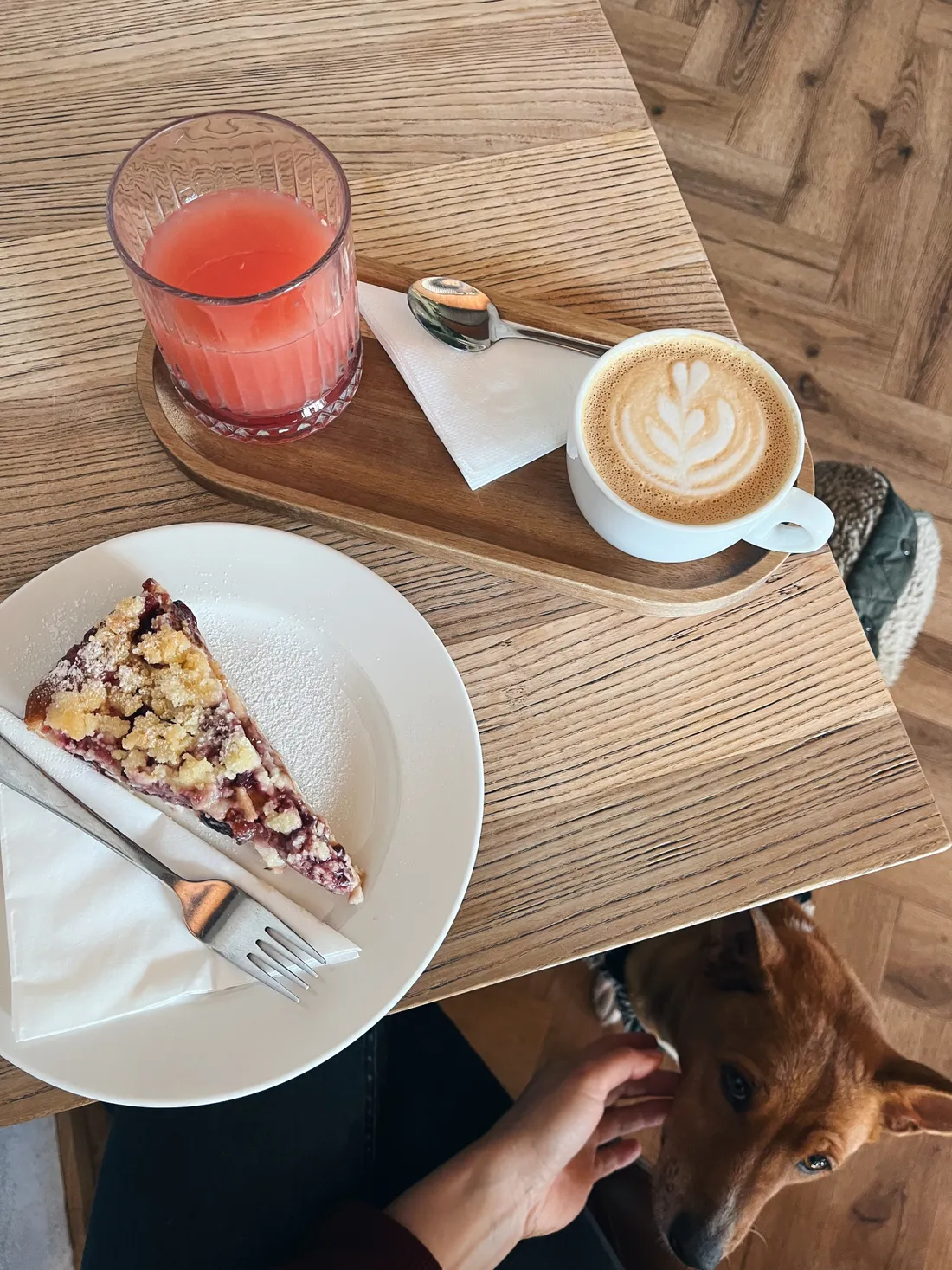 Řez ovocného koláče s drobenkou, cappuccino na dřevěném podnosu a ruka cupající psa u stolu.