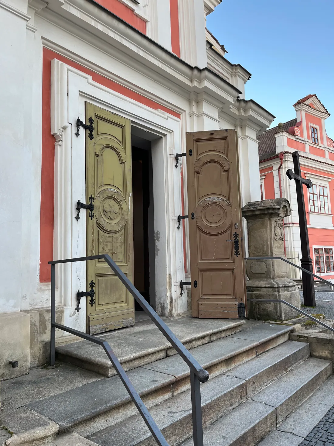 Otevřené dřevěné dveře kostela se schody, kamenným křížem a červenobílou fasádou.
