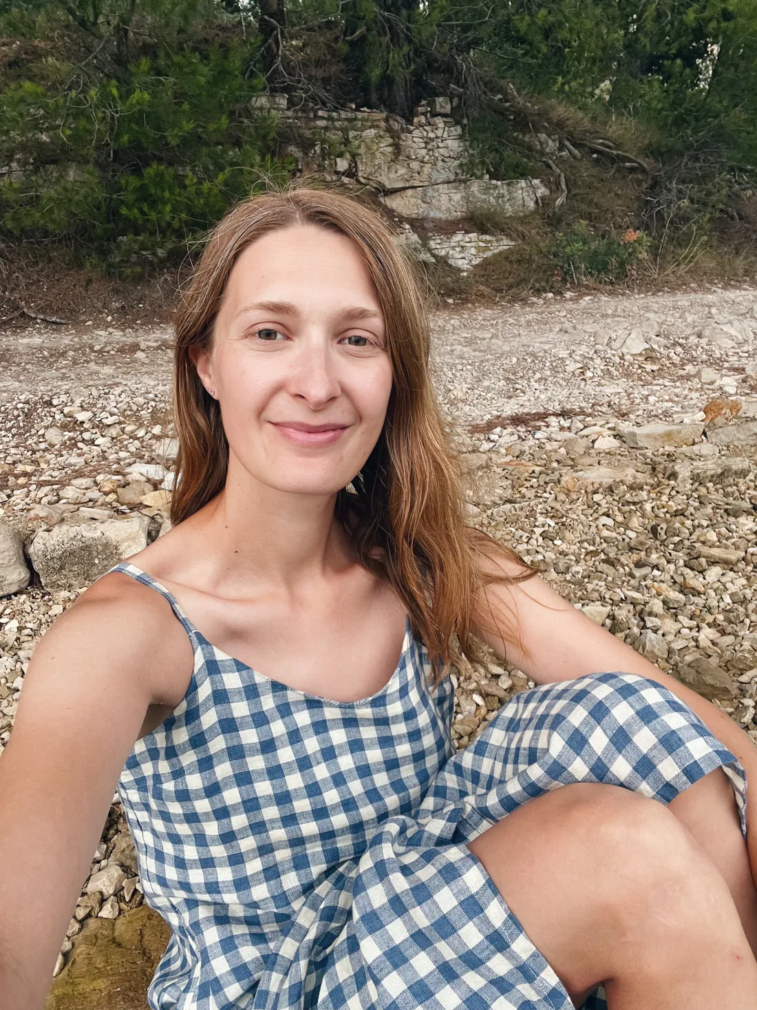 Usmívající se žena v modrobílých kostkovaných šatech sedí na oblázcích před skalnatým břehem a stromy.