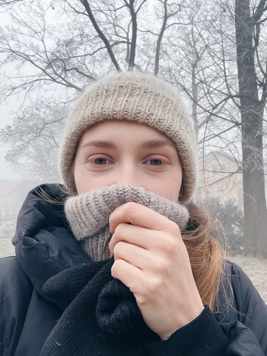 Žena v kulichu a nákrčníku v mlze, ruka v rukavici upravuje šálu u obličeje.
