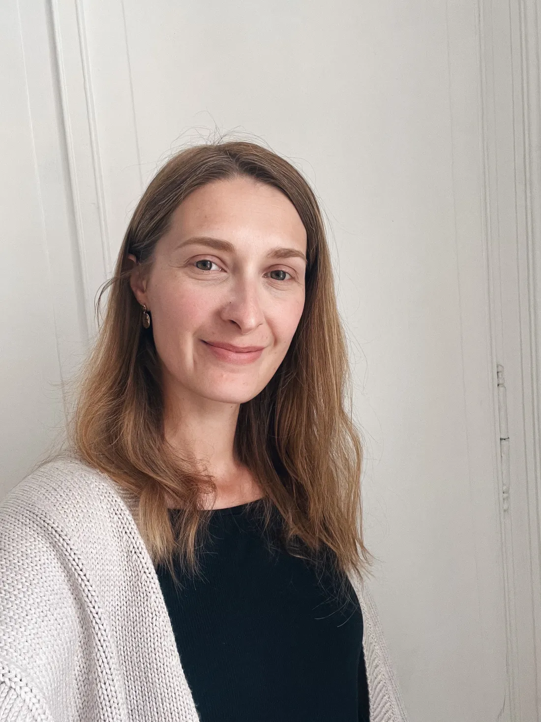 Žena se světle hnědými vlasy v šedém pleteném kardiganu se jemně usmívá před světlou stěnou.