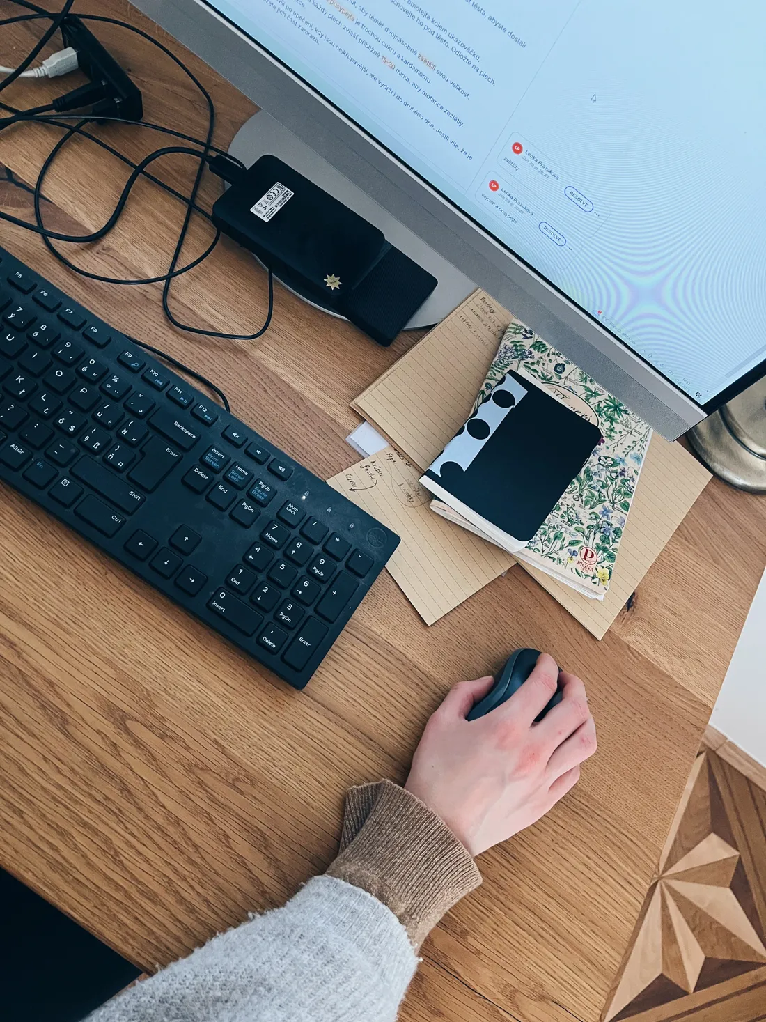 Ruka na myši u klávesnice a monitoru na světlém dřevěném stole s notebooky a kabely.