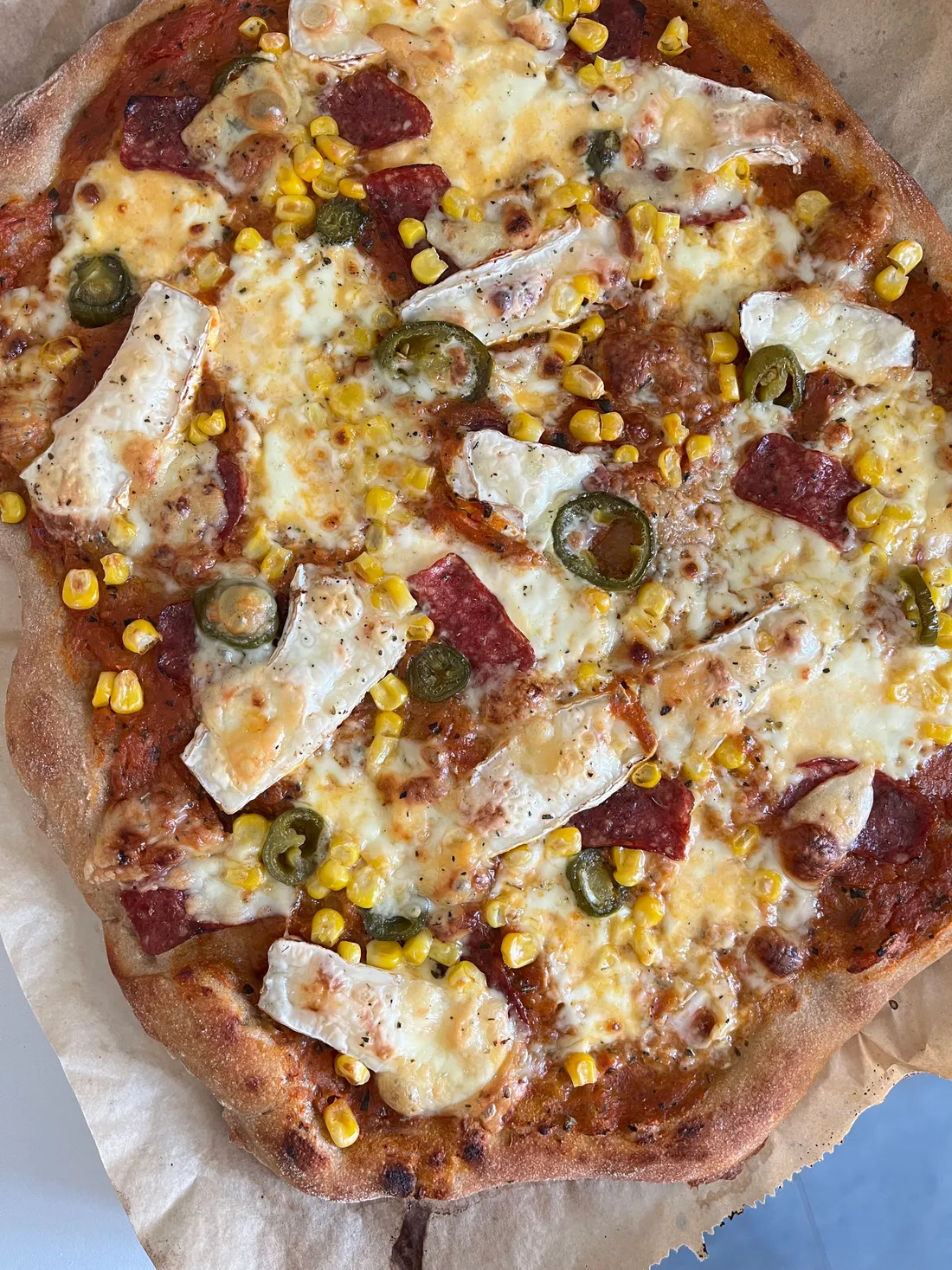 Pizza s camembertem, salámem, kukuřicí a jalapeños na papíru na světlém podkladu.