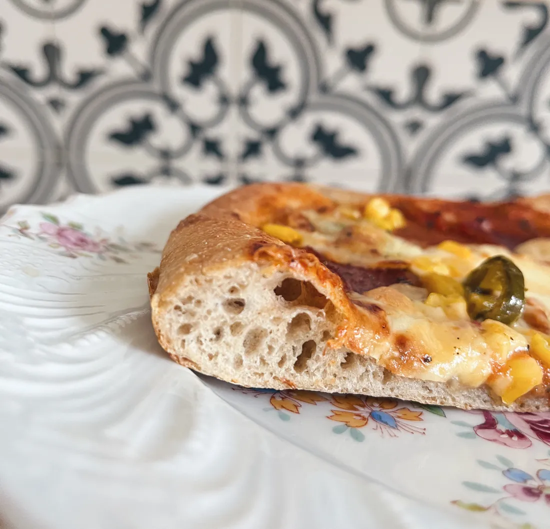 Detail nadýchaného okraje pizzy s kukuřicí a jalapeño na květovaném talíři před černobílou vzorovanou stěnou.