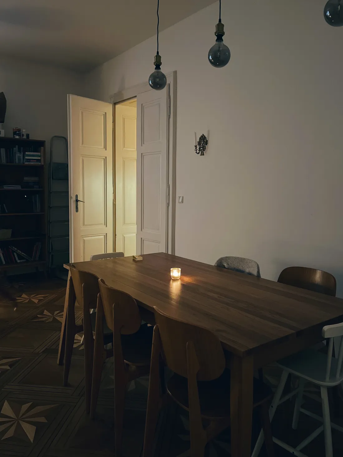 Tmavá jídelna s velkým dřevěným stolem, svíčkou a závěsnými skleněnými lampami, v pozadí otevřené bílé dveře.