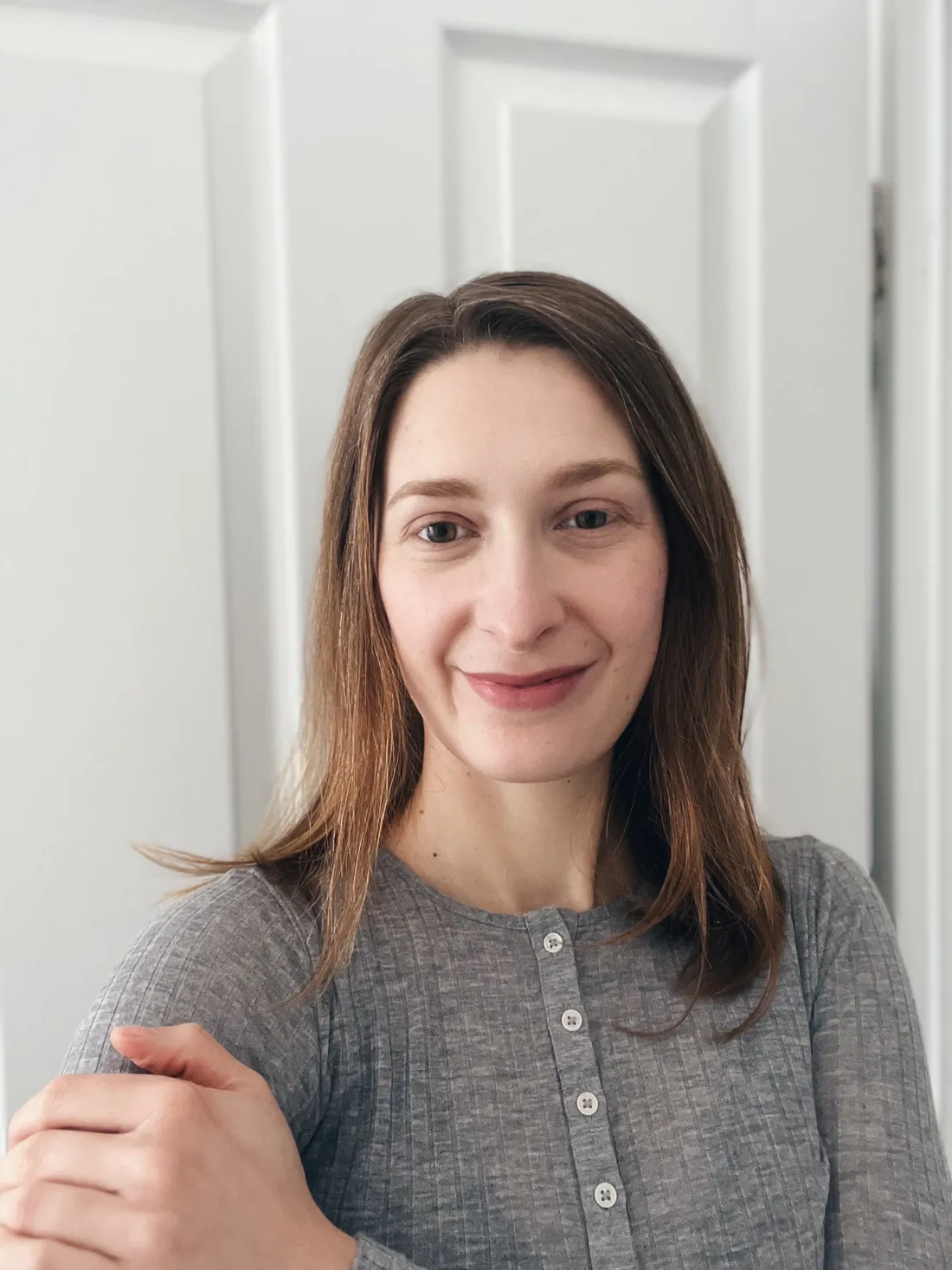 Mladá žena s hnědými vlasy a jemným úsměvem v šedém žebrovaném tričku s knoflíky před bílými dveřmi.
