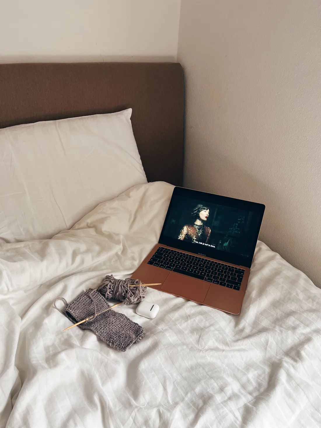Růžový notebook, šedé pletení na jehlících a bílý obal sluchátek na bílém prostěradle v posteli.