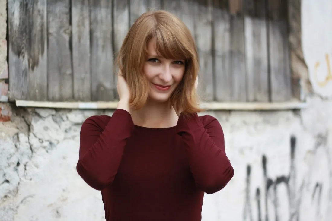 Usměvavá žena s bobem a ofinou v bordó tričku před dřevěnou a omítnutou zdí.