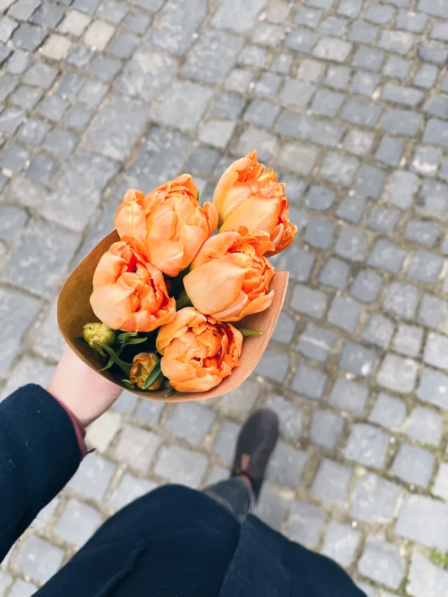 Kytice oranžových tulipánů v papíru držená nad dlažebními kostkami při procházce.