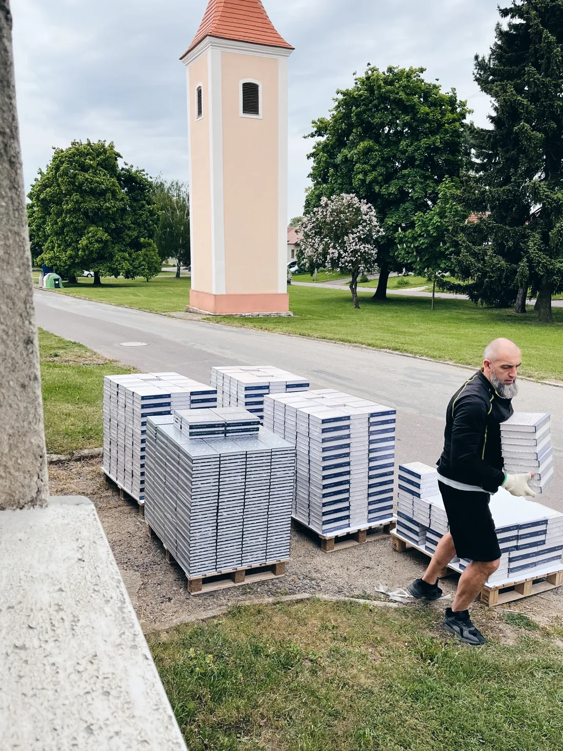 Muž skládá stohy knih z dřevěných palet venku, v pozadí vysoká věž s červenou střechou a stromy.