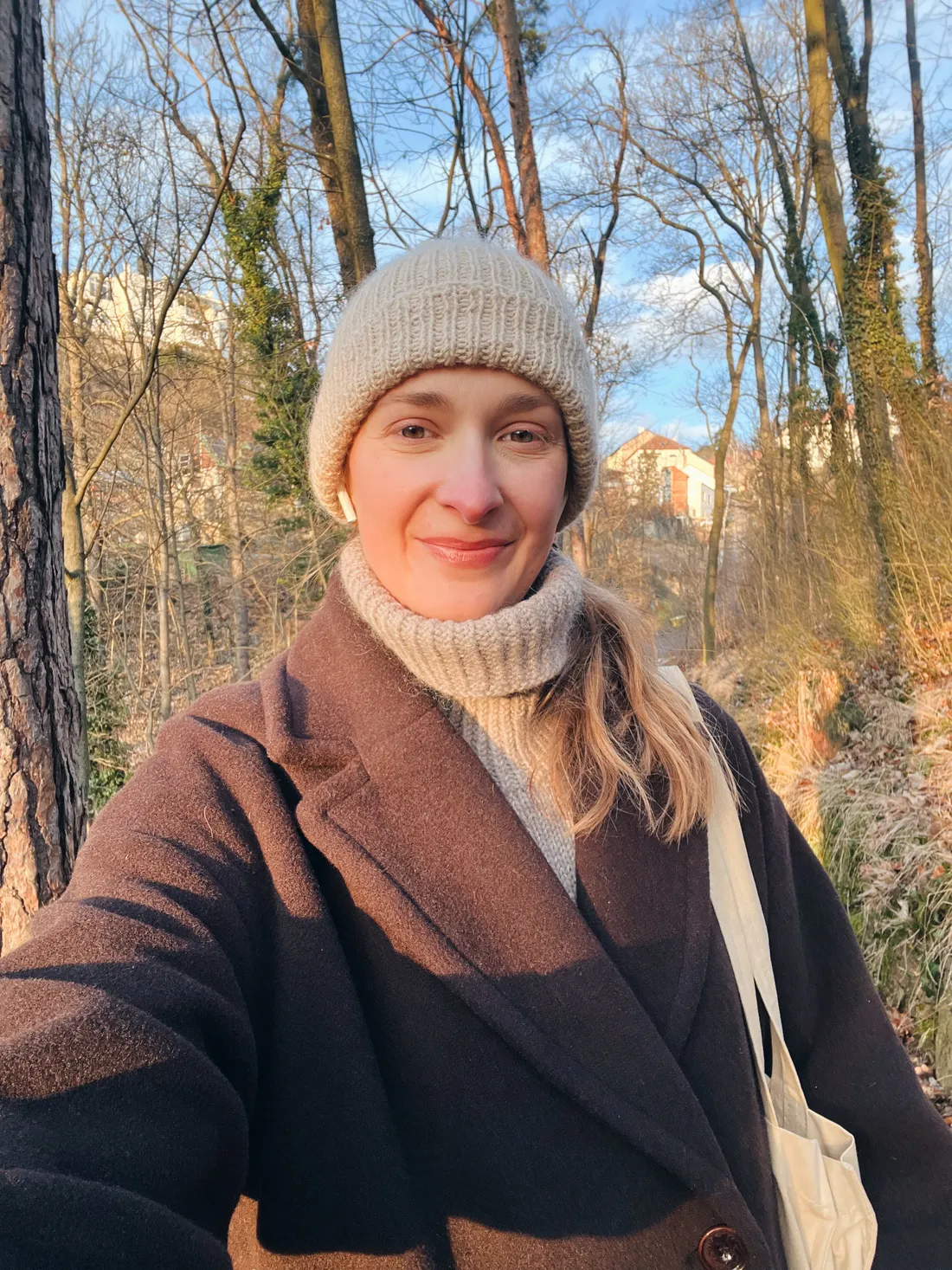 Usmívající se žena v krémové čepici a hnědém kabátu v parku při západu slunce.