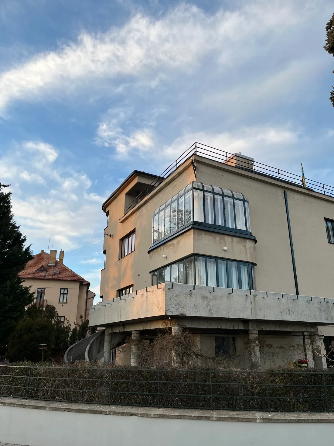 Funkcionalistická vila s výraznými prosklenými nárožními arkýři a betonovou terasou pod modrou oblohou.