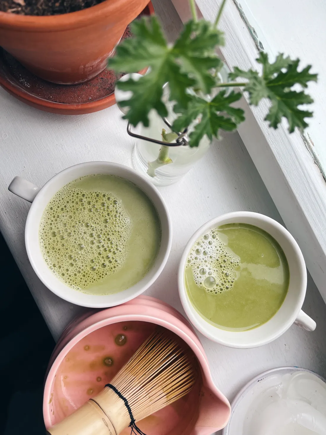 Dva šálky zeleného matcha latte na bílém parapetu s růžovou miskou a bambusovou metličkou.