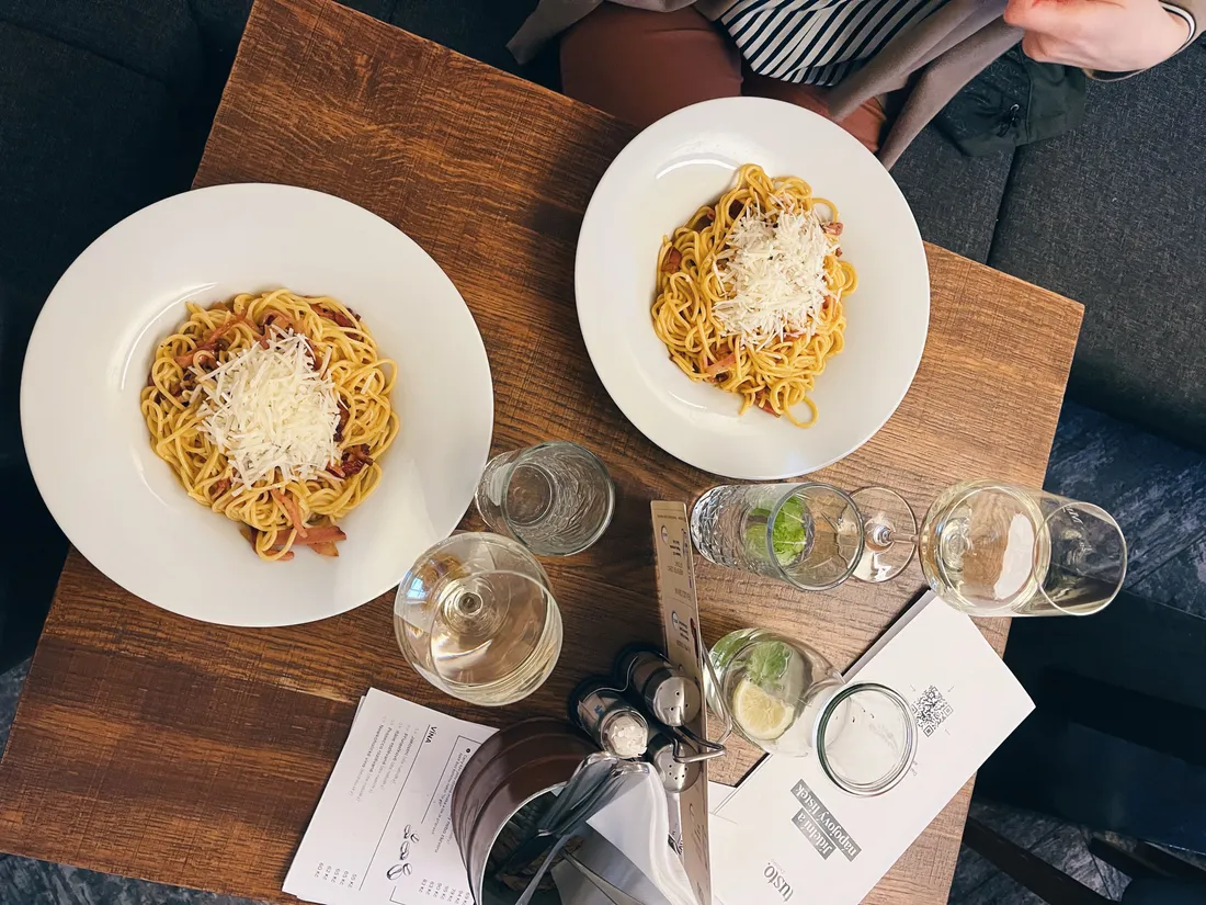 Dva talíře špaget carbonara se strouhaným sýrem a sklenice vína a vody s mátou na dřevěném stole.