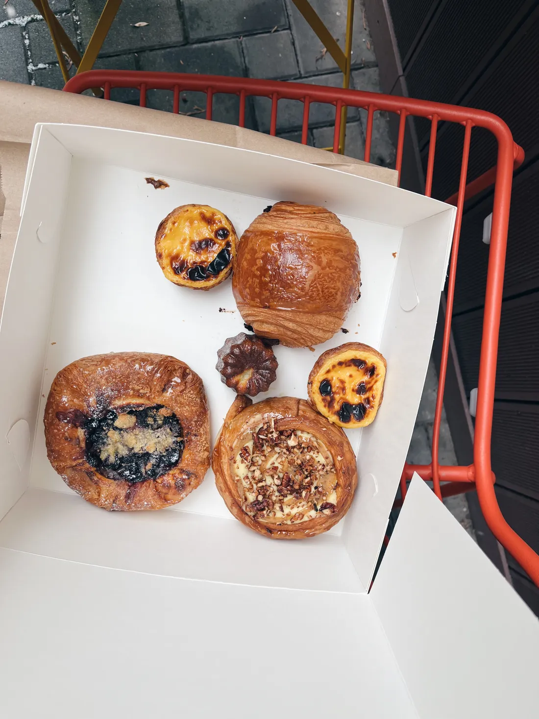 Kartonová krabička se šesti druhy pečiva — koláčky, croissant, canelé a dánské koláče na oranžové židli.
