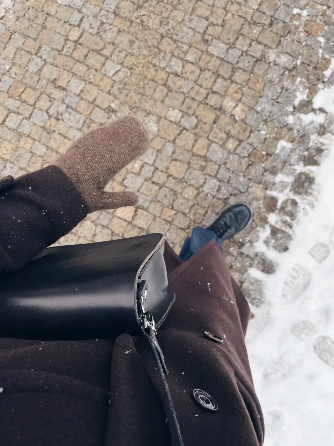 Hnědý kabát a pletená rukavice na zasněžené dlážbě, černá kožená kabelka u boku.