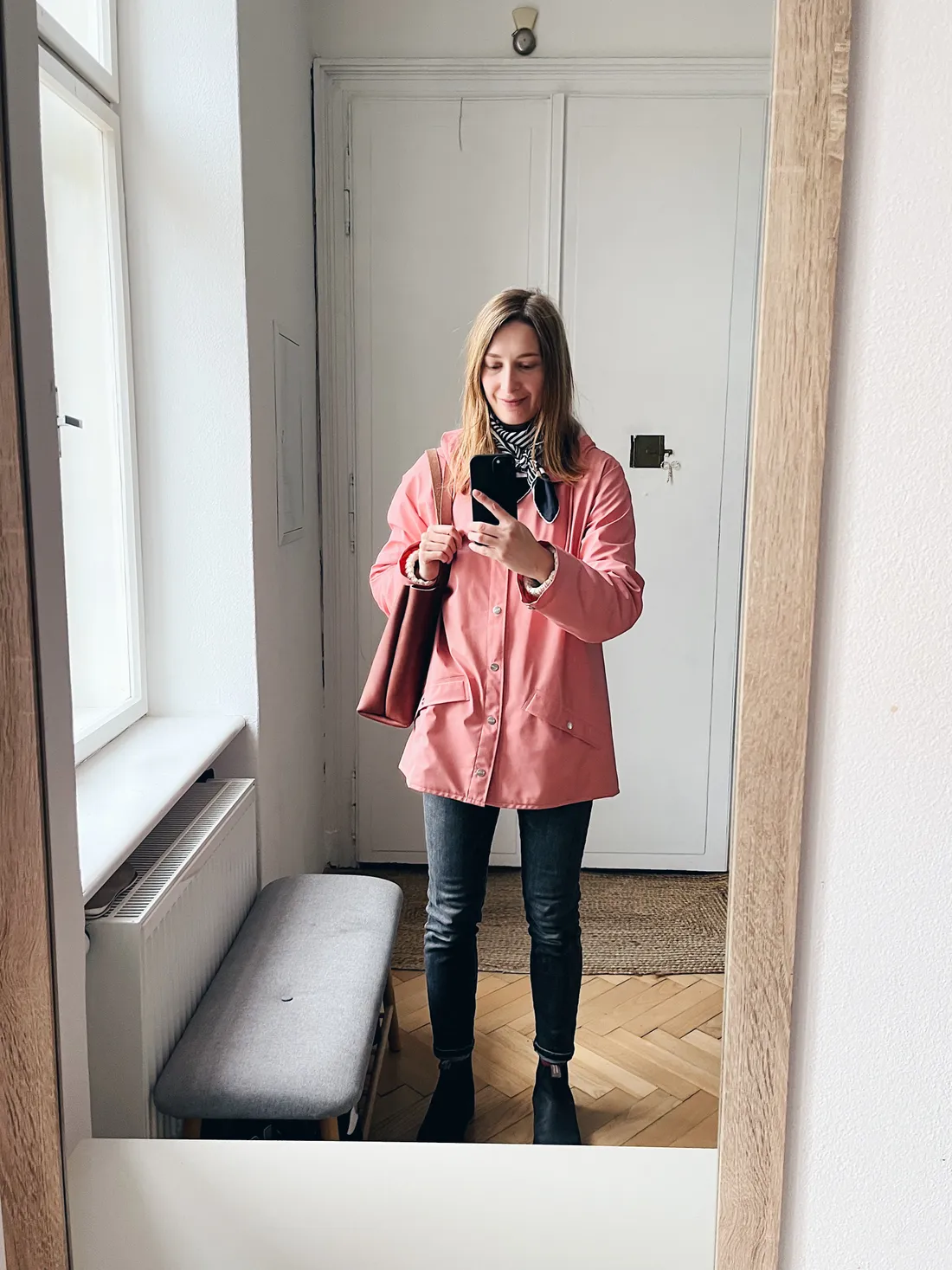 Žena v růžové pláštěnce a pruhovaném šále v zrcadlové selfie v předsíni.