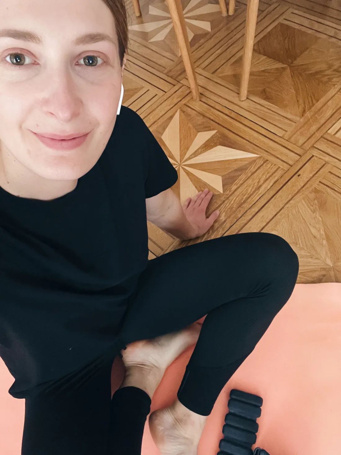 Žena v černém sportovním oblečení na růžové jógamatce s černým závažím na parketách.