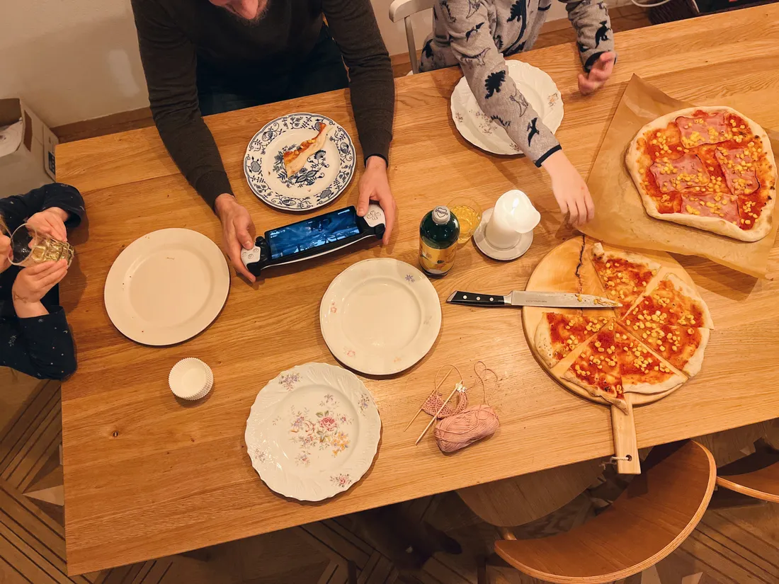 Rodina u dřevěného stolu s domácí pizzou, talíře, pití, klubíčko vlny a herní ovladač v ruce.