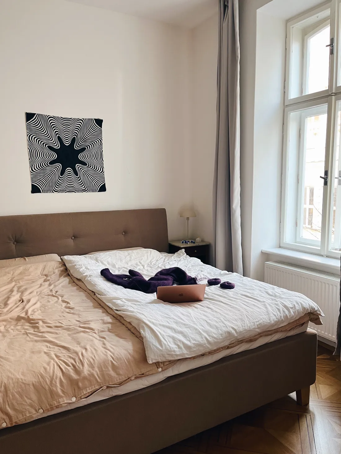 Ložnice s hnědou postelí, fialovým svetrem a notebookem, nad postelí černobílý obraz s optickým klamem.