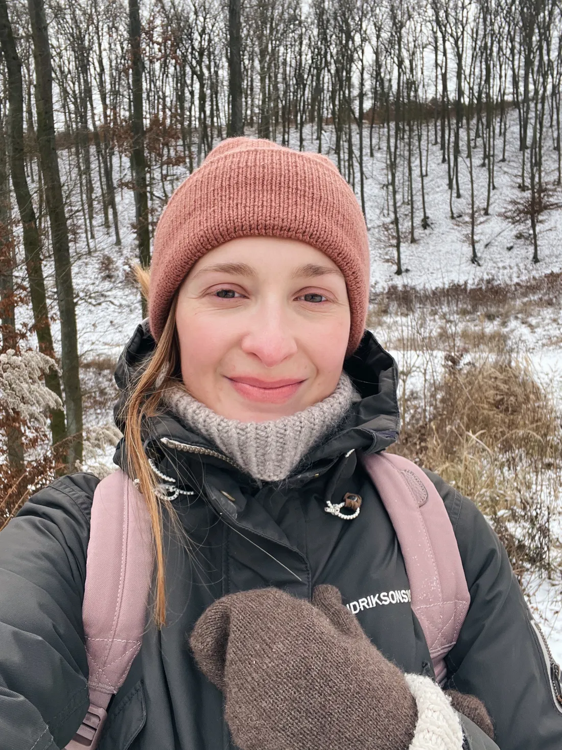 Žena v růžové čepici a tmavé zimní bundě na procházce ve sněhu, v pozadí les bez listí.