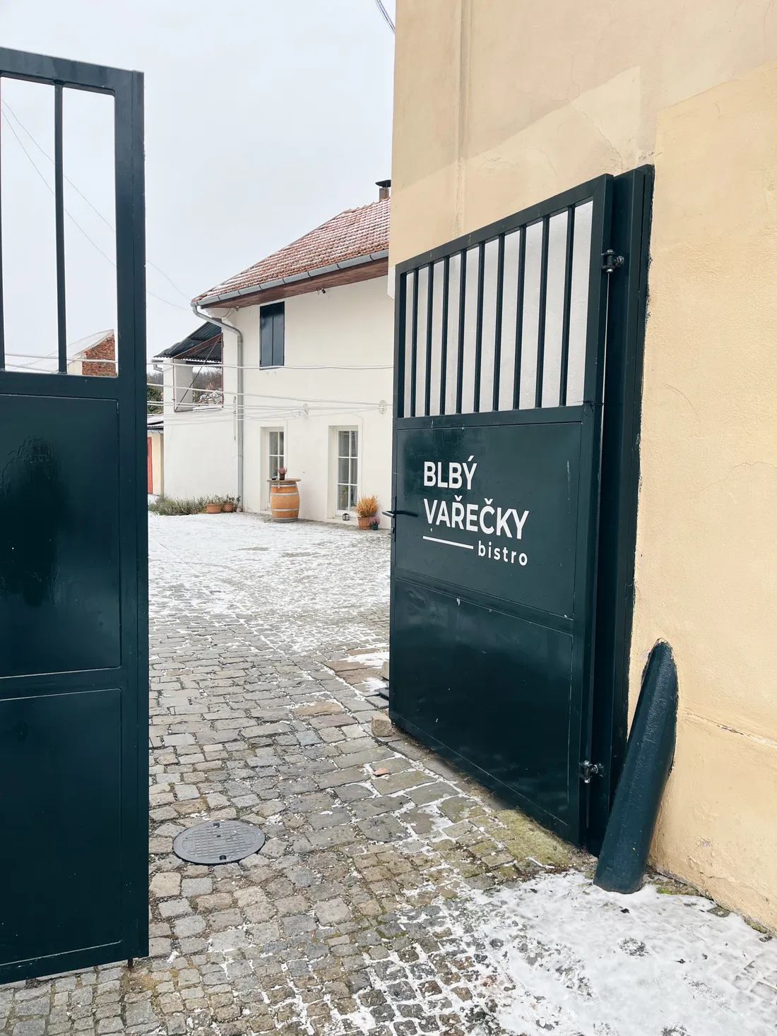 Otevřená tmavě zelená brána s nápisem Blbý vařečky bistro, dlážděný dvůr se sněhem a bílý dům.
