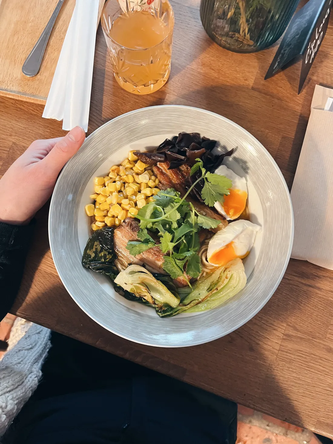 Miska ramen s bůčkem, kukuřicí, vejcem naměkko a koriandrem na dřevěném stole, ruka drží okraj.