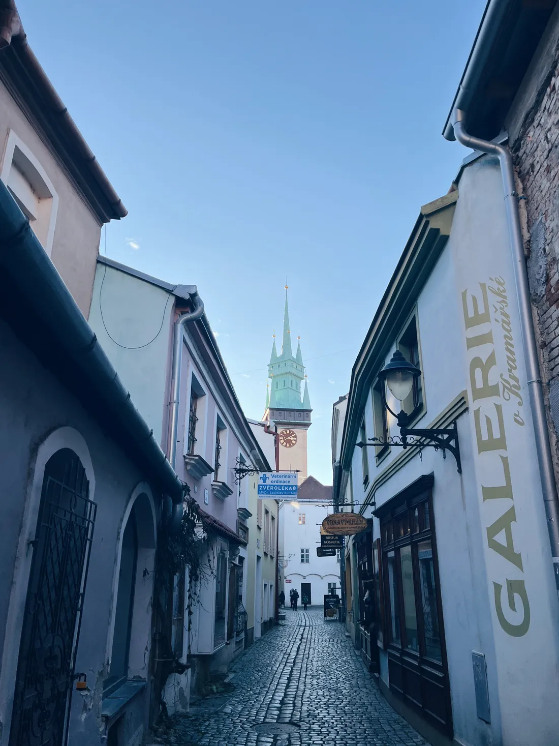 Úzká dlážděná ulička mezi historickými domy k věži se zelenou střechou a hodinami, modrá obloha.