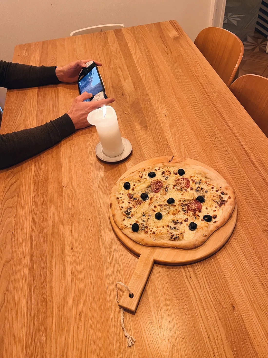 Pizza s olivami a salámem na dřevěném prkénku vedle hořící svíčky, ruka drží herní konzoli.