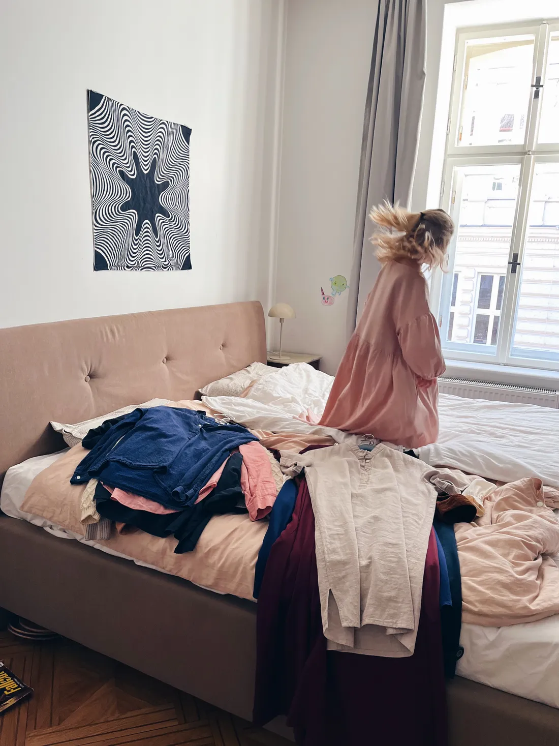 Blond dívka v růžových šatech skáče na posteli s hromadou oblečení, nad postelí černobílý plakát.