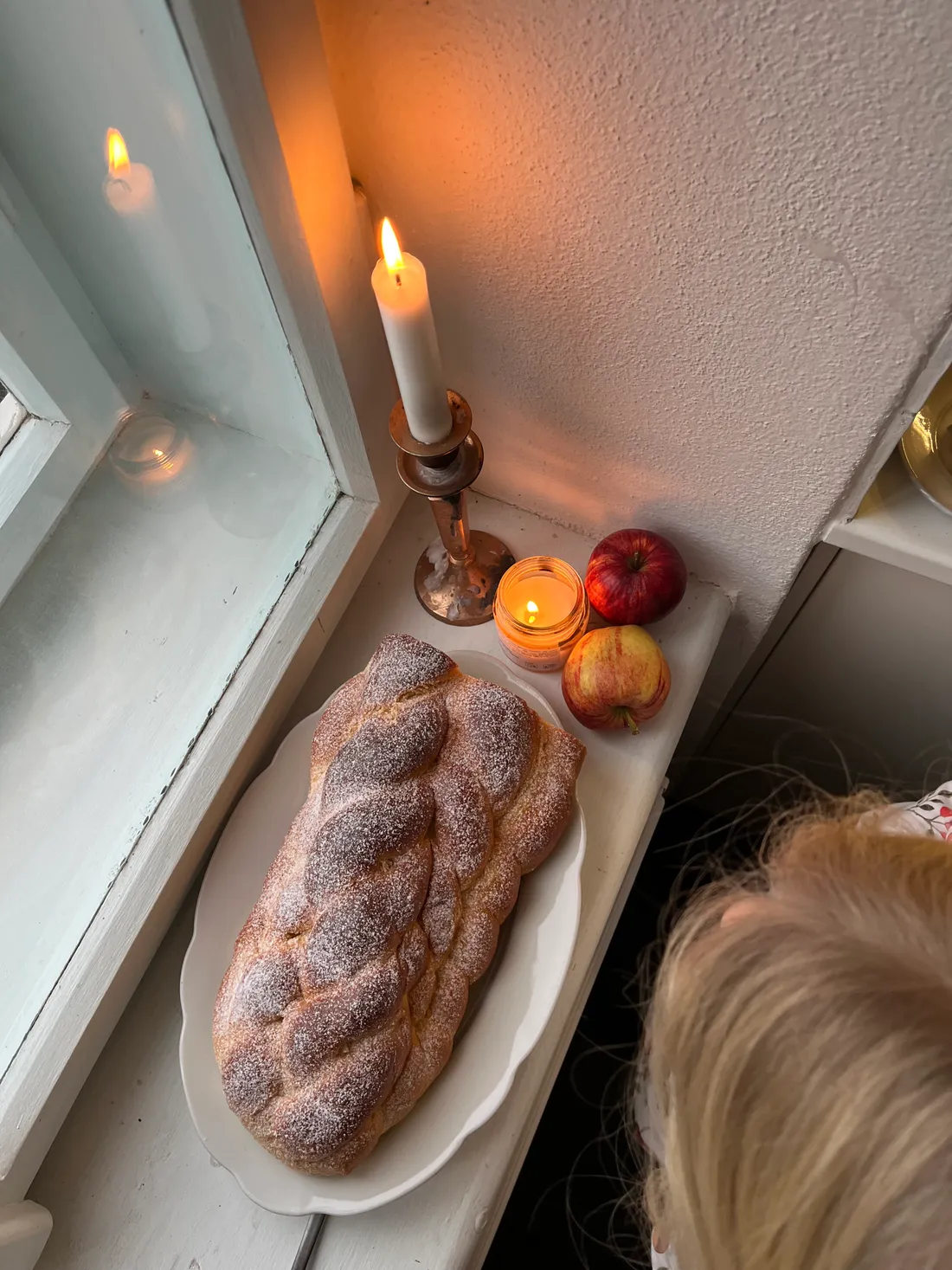 Vánočka posypaná moučkovým cukrem na bílém talíři na parapetu vedle jablek a hořících svíček.