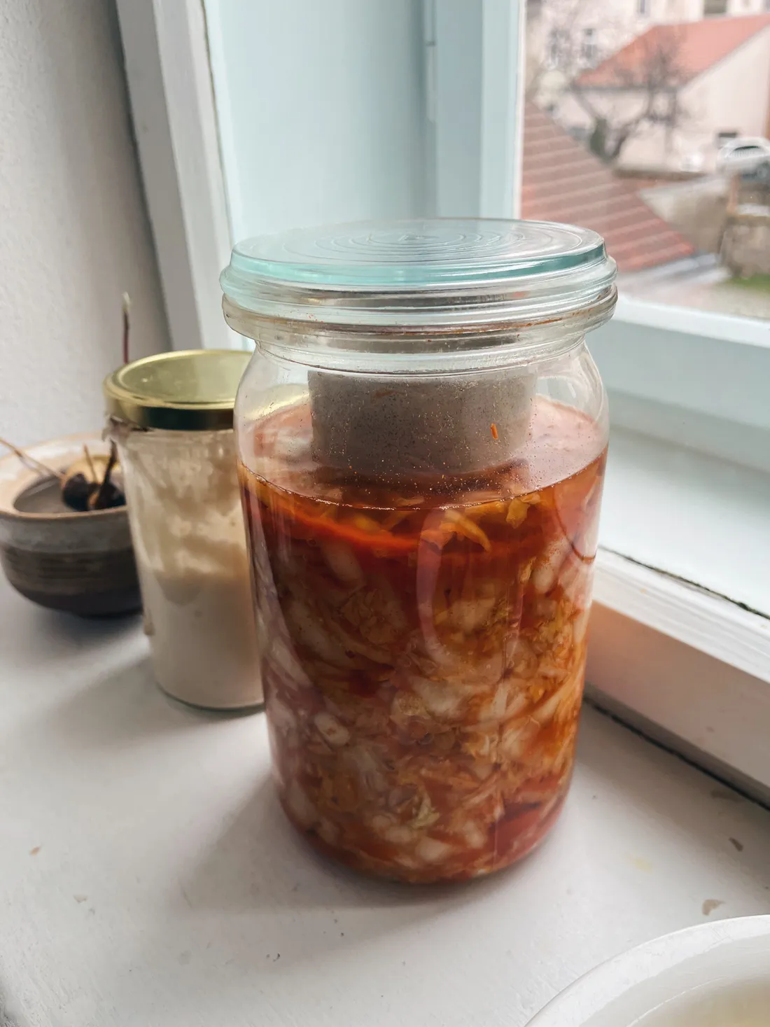 Velká sklenice kimchi s červenou omáčkou a fermentačním závažím na bílém okenním parapetu.