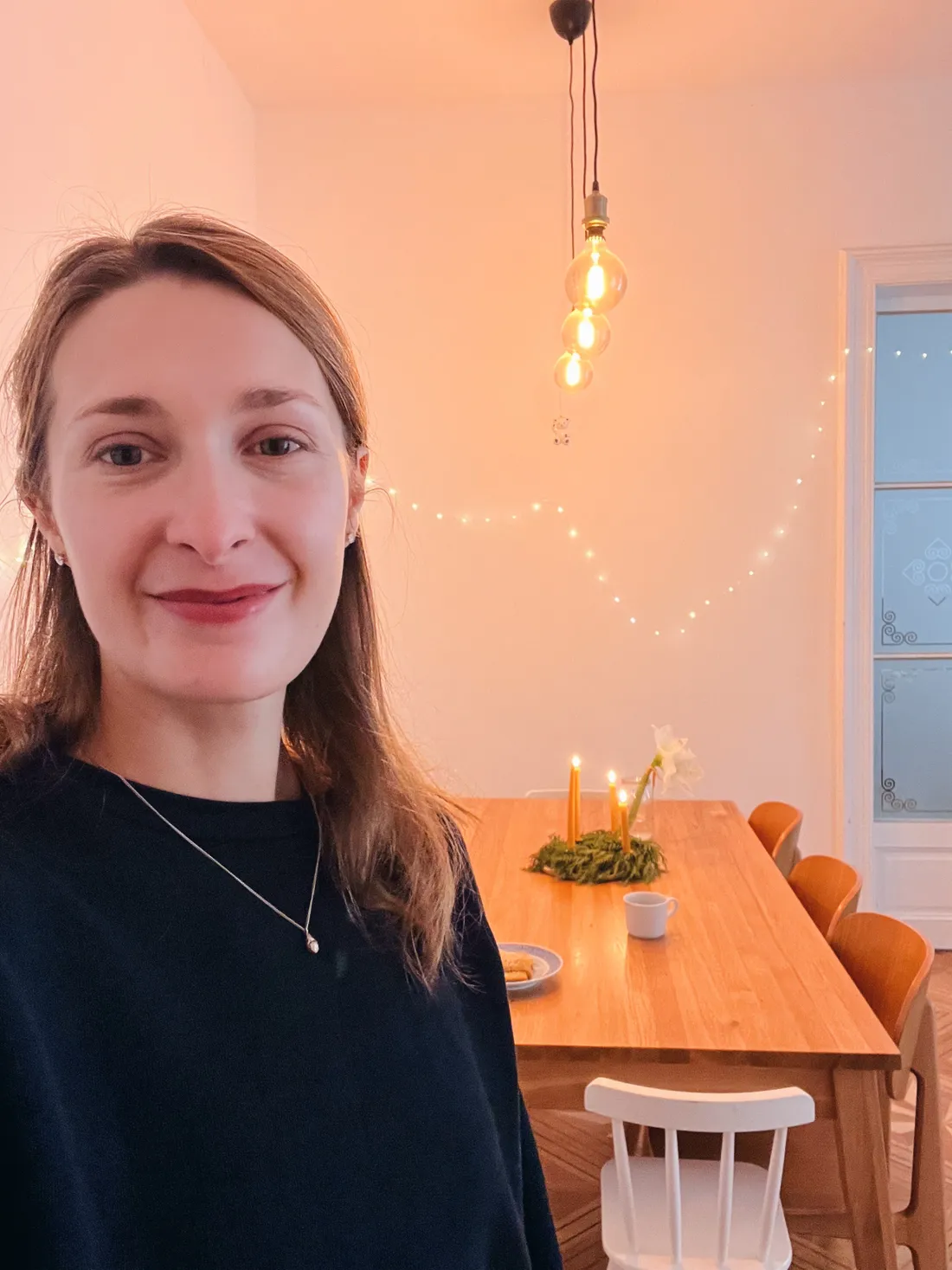 Usmívající se žena v tmavém svetru před dřevěným stolem s adventním věncem a zapálenými svíčkami.