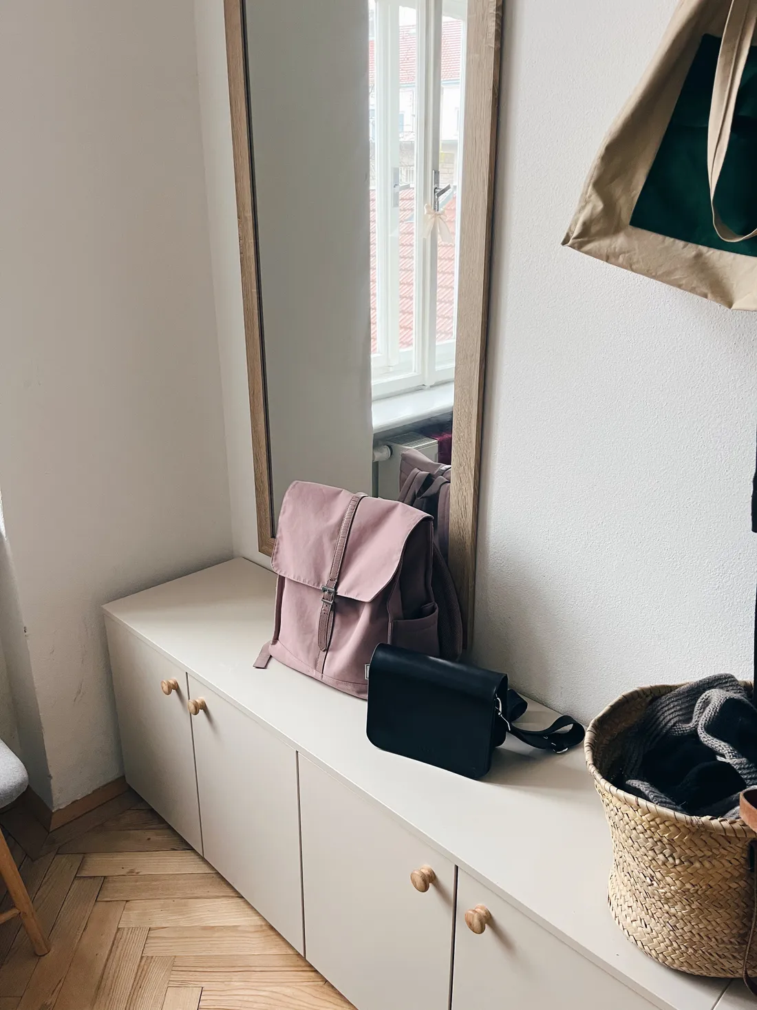 Nízká skříňka s růžovým batohlem, černou kabelkou a proutěným košem, vedle zrcadla v chodbě.